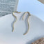 Gold asymmetrical S-curve earrings with a diamond accent on a white textured background.